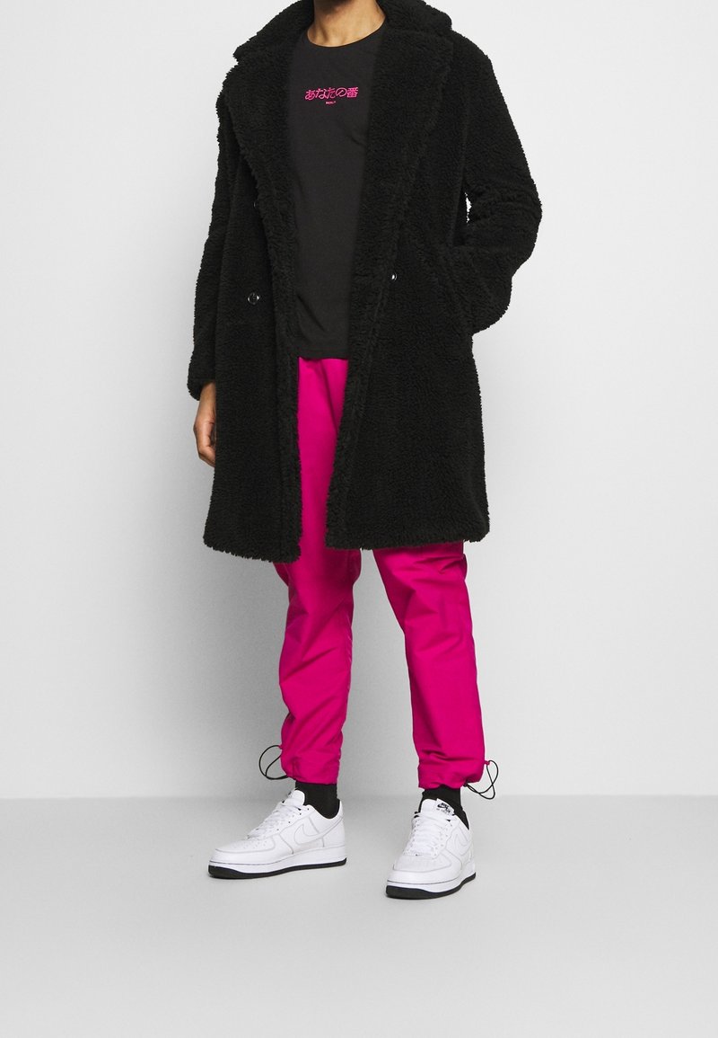 Manteau teddy noir oversize, pantalons tapered rose et baskets blanches. Le manteau présente un tissu texturé, avec un col cranté et une fermeture à bouton.