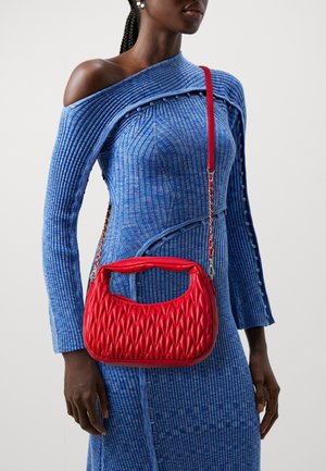 Sac à main matelassé rouge avec une anse courbée et une bandoulière en chaîne, présentant une surface texturée et une forme compacte. Porté avec une robe bleue côtelée.