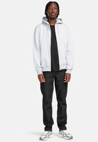 Felpa grigia con zip e cordino, indossata sopra una t-shirt nera, abbinata a jeans scuri e scarpe sportive con dettagli riflettenti.