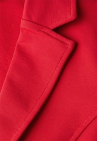 Röd tyg med en texturerad yta, utrustad med en skräddarsydd krage med kontrasterande piping och synliga sömnadsdetaljer.