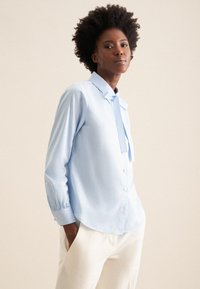 Himmelbluse mit Streifenmuster, Knopfleiste in der Front, Kragen und Nackenbindendetail; verfügt über lange Ärmel und einen abgerundeten Saum.