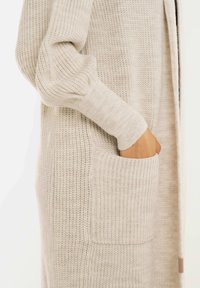 Hand in de zak van een beige gebreide cardigan met ribboordmanchetten en een gestructureerd patroon op de mouwen en het lichaam.
