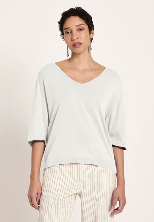 Woman wearing a loose white V-neck top with elbow-length sleeves and cream pants with thin vertical stripes, standing against a plain background.