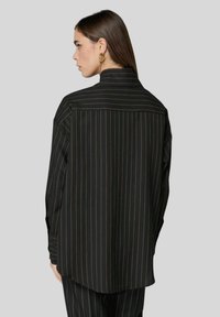 Camicia a righe nere con linee verticali dorate, maniche lunghe, yoke posteriore e orlo curvo, realizzata in tessuto morbido.