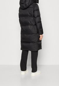 Černá péřová bunda s kapucí, charakterizovaná texturovaným povrchem, horizontálním prošitím a dlouhým, volným střihem, doplněná tmavými kalhotami a bílými teniskami.