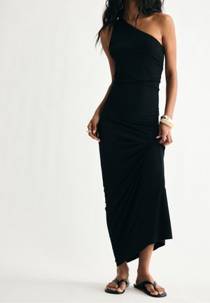 Femme portant une robe noire à une épaule, tenant l'ourlet, accessoirisée de bracelets, bagues et sandales plates noires, sur un fond uni.