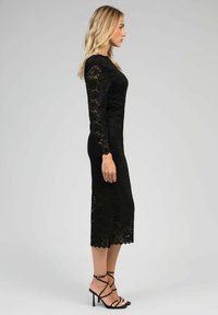 Robe en dentelle noire à manches longues, silhouette ajustée, ourlet festonné et longueur au genou. Assortie à des talons noirs à lanières.
