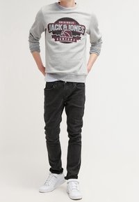 Sudadera gris con gráfico de "Jack & Jones" en burdeos oscuro; combinada con jeans negros y zapatillas blancas. Tejido suave y diseño ajustado.