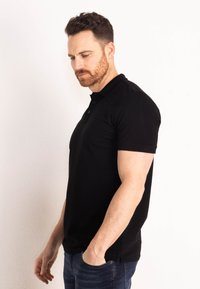 Černé polo tričko s krátkými rukávy, klasickým límcem a třemi předními knoflíky. Nosené s modrými džínami. Hladká textura a přiléhavý střih.
