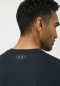 Black athletic shirt featuring a round neckline, smooth texture, and a gray Under Armour logo at the back near the collar.