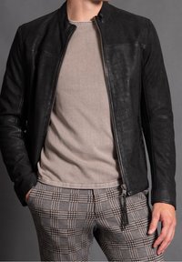 Svart läderjacka med dragkedja framtill och utan krage, kombinerad med en ljusbeige skjorta och rutiga byxor.