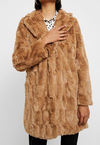 Fuskpälsjacka i ljust brunt med en lurvig textur. Har en sjalkrage och vida ärmar, bärs över en mönstrad skjorta.