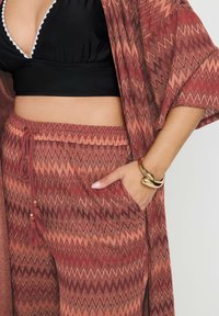 Bordeauxfärgade byxor med zickzackmönster och dragsko i midjan, tillsammans med en matchande kimonotäckning. Svart kort topp under, guldarmband på handleden.