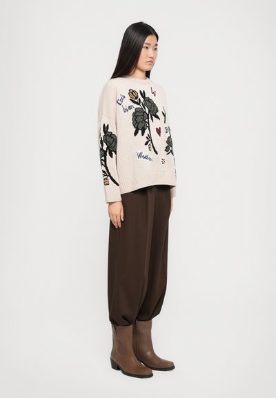Gebreide trui in lichtroze, met bloemige borduursels in zwart en kleuraccenten. Bruine broek met brede pijpen en bruine enkellaarzen maken de look compleet.