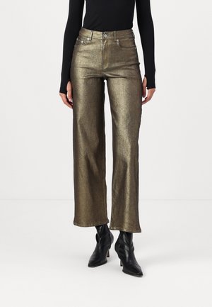 Personne portant un pantalon doré métallique droit, un haut noir à manches longues et des bottines noires à talons sur fond blanc.