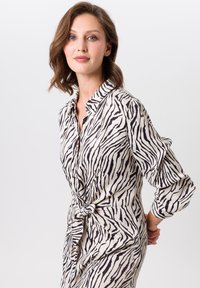 Robe chemise à manches longues en crème avec des rayures zèbre noires, présentant une fermeture à boutons sur le devant et un détail de taille nouée. Texture de tissu lisse.
