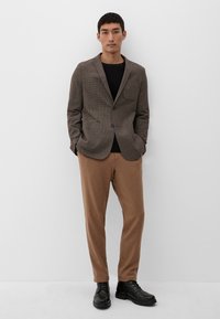 Braun kariertes Sakko mit einer Brusttasche, kombiniert mit beigen Hosen und schwarzen Schnürstiefeln. Das Modell steht vor einem schlichten Hintergrund.