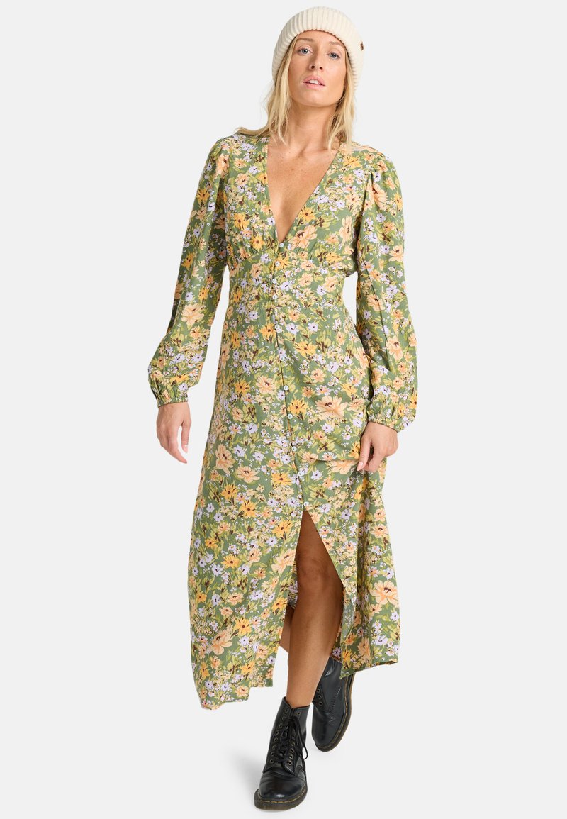 Vestido maxi floral en verde con flores amarillas y moradas. Presenta mangas largas abullonadas, un escote en V profundo, cierre frontal de botones y una apertura lateral.