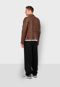 Giacca di pelle marrone con colletto rialzato, tessitura liscia e dettagli di cucitura, abbinata a pantaloni neri a gamba larga e sneakers.