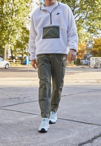 Valkoinen fleece-pullover vihreällä vetoketjutaskulla, yhdistetty oliivinvärisiin cargo-housuihin, joissa on sivutaskut, sekä valkoisiin lenkkareihin pastelliväreillä.