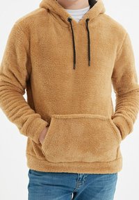 Beżowa bluza z kapturem wykonana z miękkiego materiału typu polar, z regulowanym sznurkiem przy kapturze i przednią kieszenią kangurką. Posiada ściągacze na mankietach i u dołu.