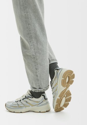 Person wearing light gray jeans, dark gray ribbed socks, and white sneakers with beige and black accents, showing the sole of one shoe.