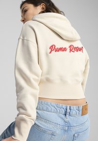 Skraćena bež dukserica s kapuljačom, sa crvenim izvezenim natpisom "Puma Resort" na leđima. Mekani materijal s rebrastim manžetama i rubom.