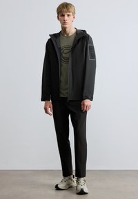 Fekete könnyű dzseki kapucnival, elöl cipzárral és oldalzsebekkel. Olívazöld póló és fekete nadrág fölött viselve. Semleges színű sportcipő.