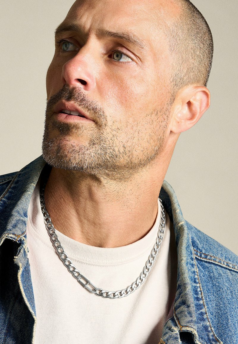 Silversmyckets kedja bärs över en ljusbeige skjorta, som kontrasterar med en denimjacka. Kedjan har tjocka länkar och en robust lås.