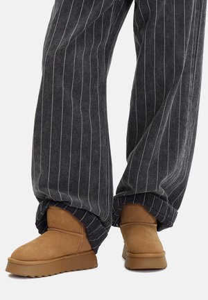 Braune Wildleder-Stiefeletten mit dicker Gummisohle, getragen zu übergroßen schwarzen Nadelstreifenhosen, mit hochgekrempelten Bündchen über den Stiefeln.