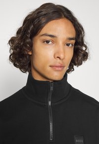 Zwarte zip-up sweatshirt met een hoge kraag, voorzien van een gladde textuur en een klein logo op de borst. Minimalistisch design en effen kleur.