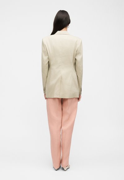 Γυναίκα όρθια, γυρισμένη με την πλάτη, που φοράει μπεζ σακάκι, ροζ παντελόνια με φαρδιά μπατζάκια και μυτερά τακούνια, με φόντο απλό λευκό.