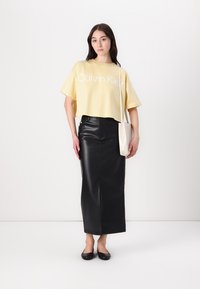 Kuttet gul t-skjorte med hvitt logo, kombinert med et høyt liv svart lær blyantskjørt og svarte leiligheter, accessorized med en lys veske.