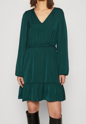 Vrouw draagt een donkergroene jurk met lange mouwen, een V-hals, een geplooide taille, een gerimpelde zoom en zwarte kniehoge laarzen tegen een effen achtergrond.