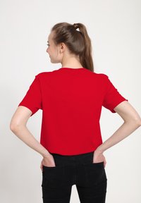 Červené tričko s krátkými rukávy vyrobené z hladkého bavlněného materiálu, s kulatým výstřihem a volným střihem, nošené s černými džíny, pohled ze zad.