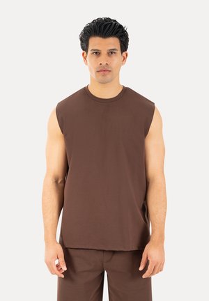 Hombre con cabello oscuro que lleva una camiseta sin mangas marrón y pantalones marrones a juego, de pie contra un fondo claro y liso.