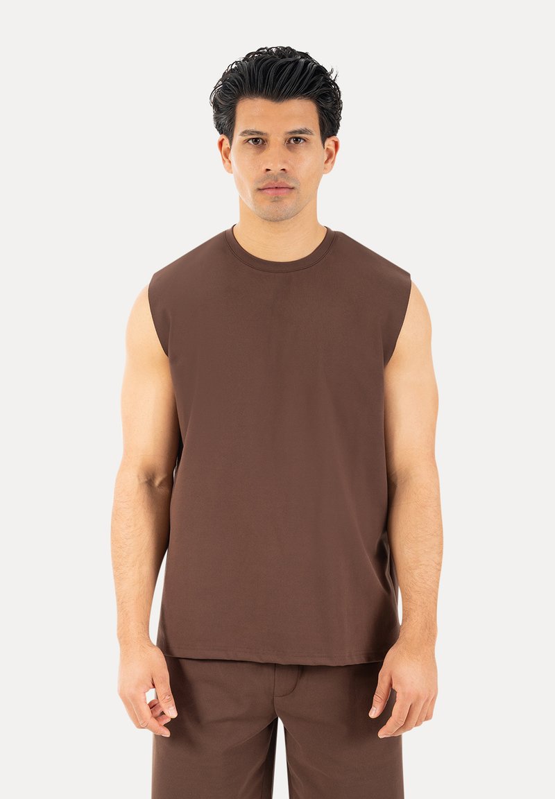 Homme aux cheveux foncés portant un déb sleeveless marron et un pantalon marron assorti, debout devant un fond clair uni.