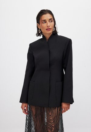Zwarte blazer met een hoge kraag, lange mouwen en twee zijzakken; vergezeld van doorschijnende zwarte patroonelementen aan de zoom.