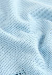 Šviesiai mėlynas tekstūruotas audinys su vaflinio pynimo raštu ir mažomis siūtomis etiketėmis, rodančiomis dalinę koordinatę.