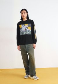 Schwarzes Langarmshirt mit einem grafischen Design in Grau, Gelb und Weiß, kombiniert mit lockeren grünen Jeans und weißen Sneakern.