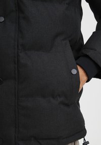 Sort isoleret jakke med en tekstureret overflade, der har en stor frontlomme sikret med en sort trykknap og elastiske manchetter omkring ærmerne.