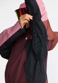 Veste isolante colorée avec des segments en noir, bordeaux et rose. Elle possède une ouverture de poche zippée sur la manche.