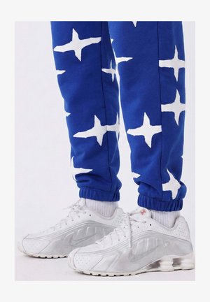 Blauwe joggingbroek met witte sterpatronen en elastische boorden, gecombineerd met witte en zilveren Nike-sneakers met geperforeerde details en veters.