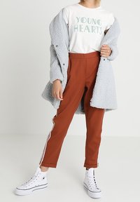 Cappotto in pelliccia sintetica grigia sopra una maglietta bianca con il testo "YOUNG HEARTS", pantaloni a gamba affusolata color ruggine con strisce laterali e sneakers alte bianche.