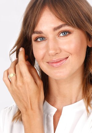 Gold ring with a smooth, circular top on a slender band, worn on a relaxed hand. Background is neutral with soft lighting.