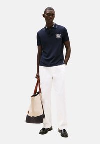 Polo de color marino con ribete blanco, logo bordado. Combinado con pantalones blancos y un bolso tote con acentos en beige claro y azul marino oscuro.