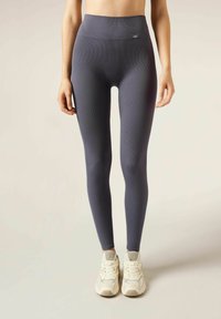 Hoch-taillierte, gerippte Leggings in einem Dunkelgrau-Ton, aus dehnbarem Stoff gefertigt. Kombiniert mit hellen Sportschuhen.