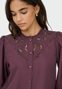 Jonge vrouw kijkt omlaag en draagt gouden creolen en een pruimenblouse met bloemenkant yoke en knopen aan de voorkant.