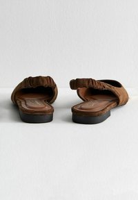 Chaussures slip-on en daim marron avec un accent froncé sur le dessus, semelle plate noire et semelle intérieure rembourrée.