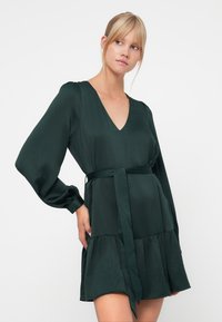 Dark green dress with a V-neck, long puffed sleeves, cinched waist with a tie, and a tiered skirt. Smooth fabric with a subtle texture.